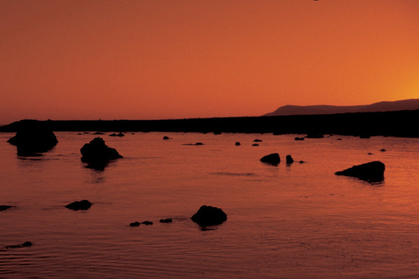 Rocky shoreline at sunset with calm water and coastal hills in the background