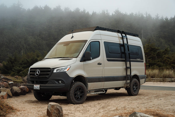 Custom Sprinter van with off-road tires and ladder parked on a sandy road by a forest