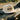 A white flat-brim cap with a black 'Sandy Vans' logo printed on the front, resting on a car dashboard with a blurred outdoor road and trees in the background.