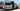 Custom white Sprinter van with open side door, roof rack, and off-road tires, parked outdoors.